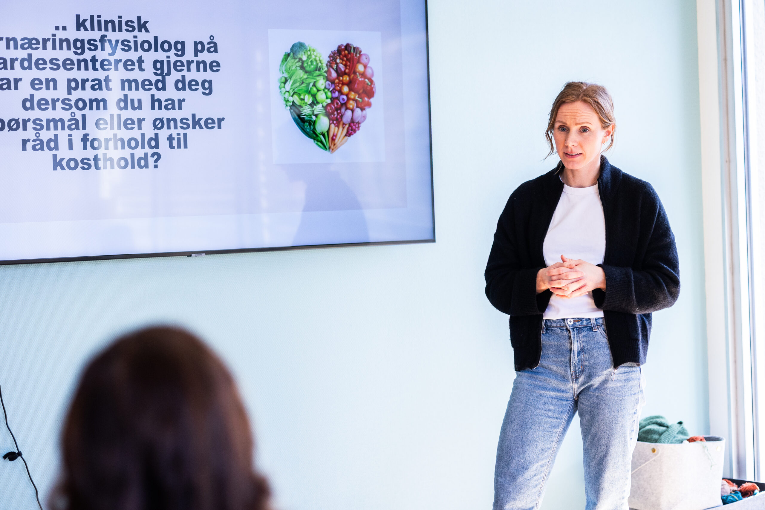 En kvinne står og snakker ved siden av en skjerm som viser norsk tekst om ernæring, med et hjerteformet bilde av ulike matvarer.