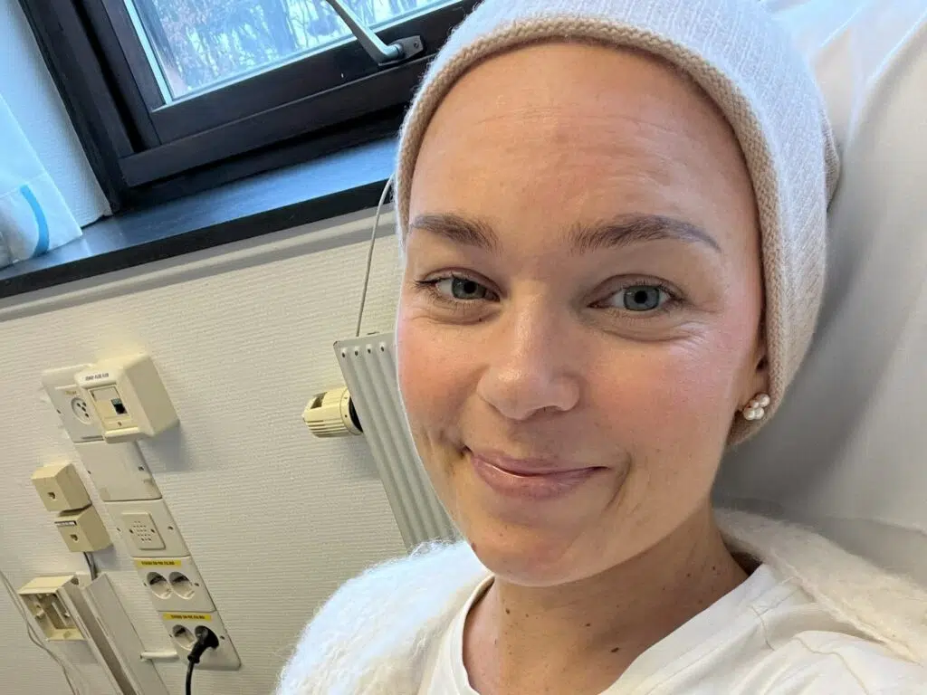 En person i hvit skjorte og lue smiler mens han sitter i et medisinsk miljø med utstyr og stikkontakter synlige i bakgrunnen.