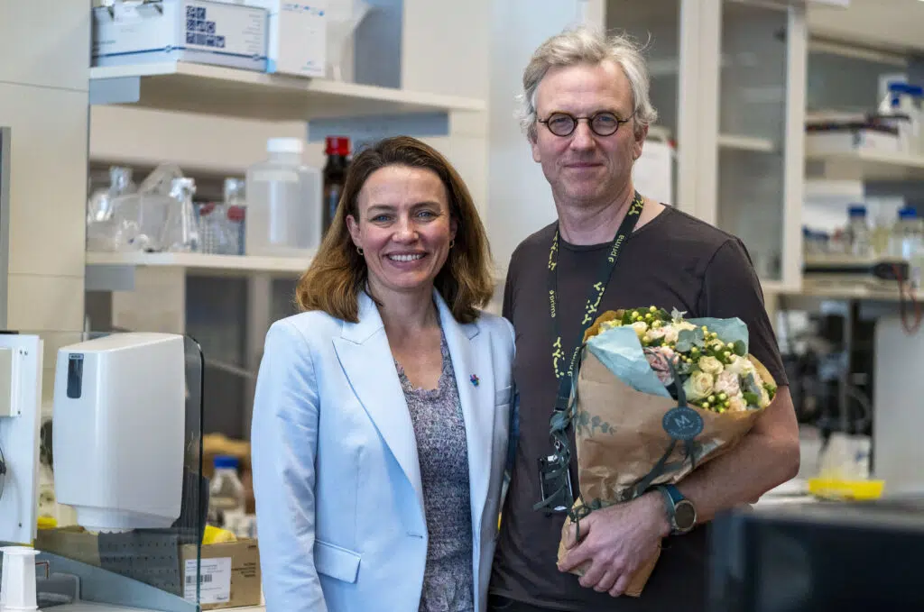 To personer står i et laboratorium; man holder en bukett blomster. Hyller med laboratorieutstyr og utstyr er synlige i bakgrunnen.