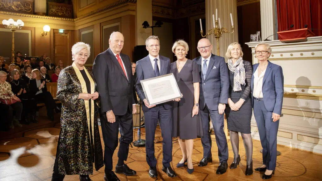 En gruppe på syv personer står sammen innendørs, med én person som har et innrammet sertifikat. De er kledd i formell antrekk, og settingen ser ut til å være en formell begivenhet eller seremoni.