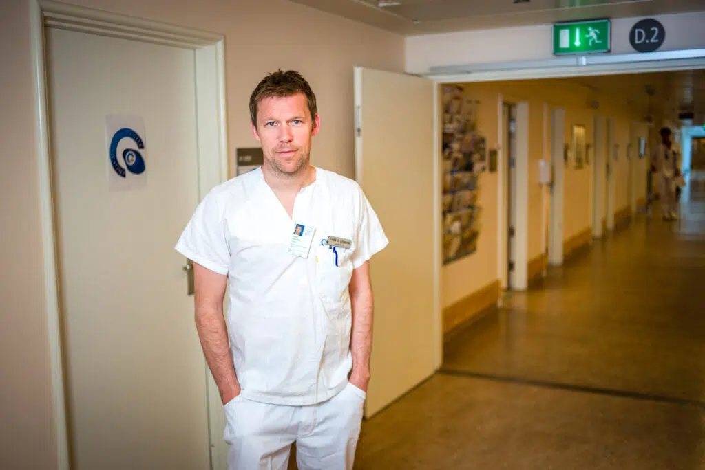 En mann i hvit medisinsk uniform står i en sykehusgang nær en dør merket med et øyesymbol, med identifikasjonsmerke og penner i lommen.