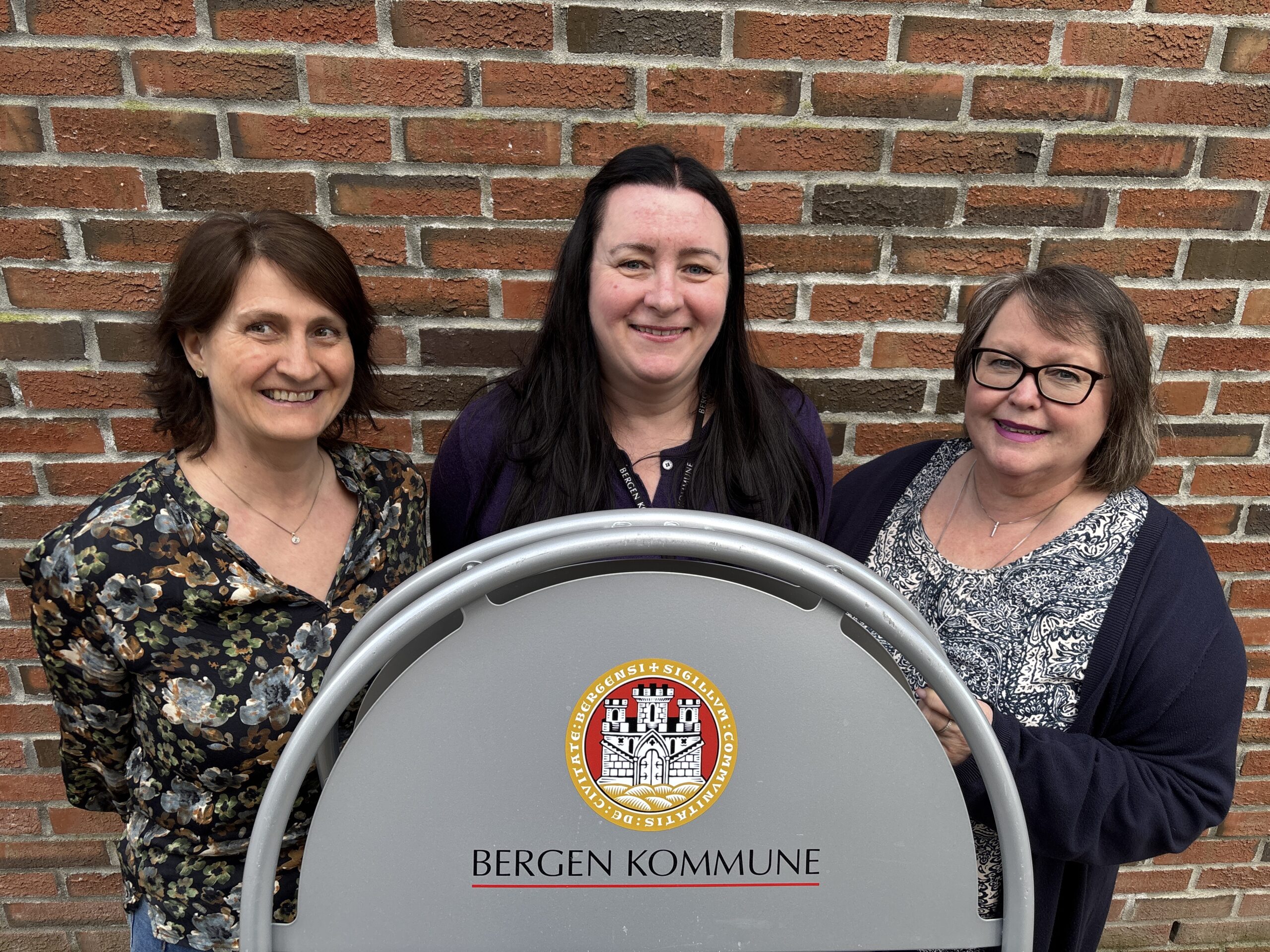 Tre kvinner står bak et skilt med Bergen Kommune-emblem, og poserer foran en murvegg.