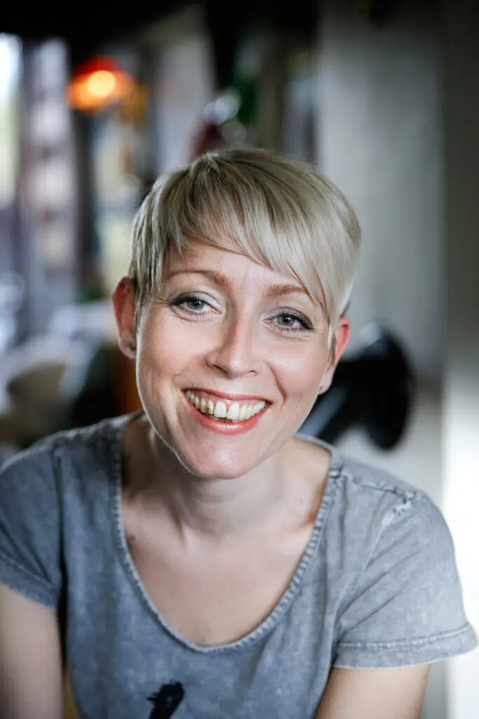 En person med kort, blondt hår og grå skjorte smiler til kameraet i en innendørs setting.