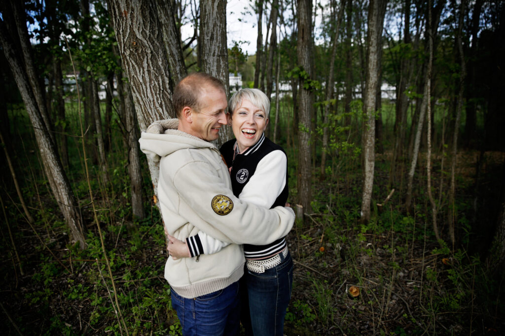 En mann og kvinne som smiler og klemmer i en skog, omgitt av trær.