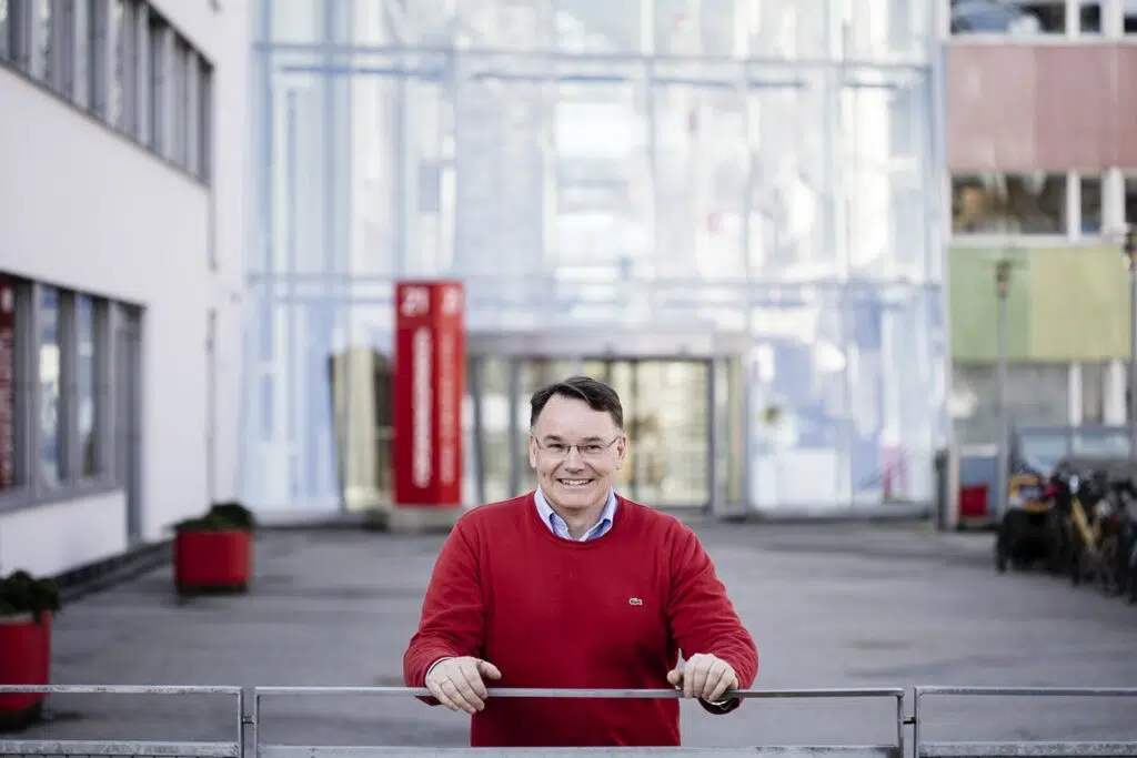 En person i rød genser står utendørs og smiler mens han lener seg på et metallrekkverk foran en moderne bygning.