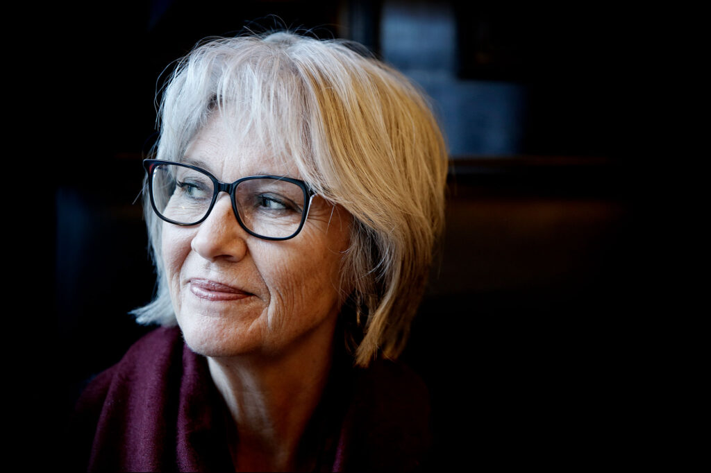 Eldre kvinne med hvitt hår og briller, iført et vinrødt skjerf, ser til siden mot en mørk bakgrunn.