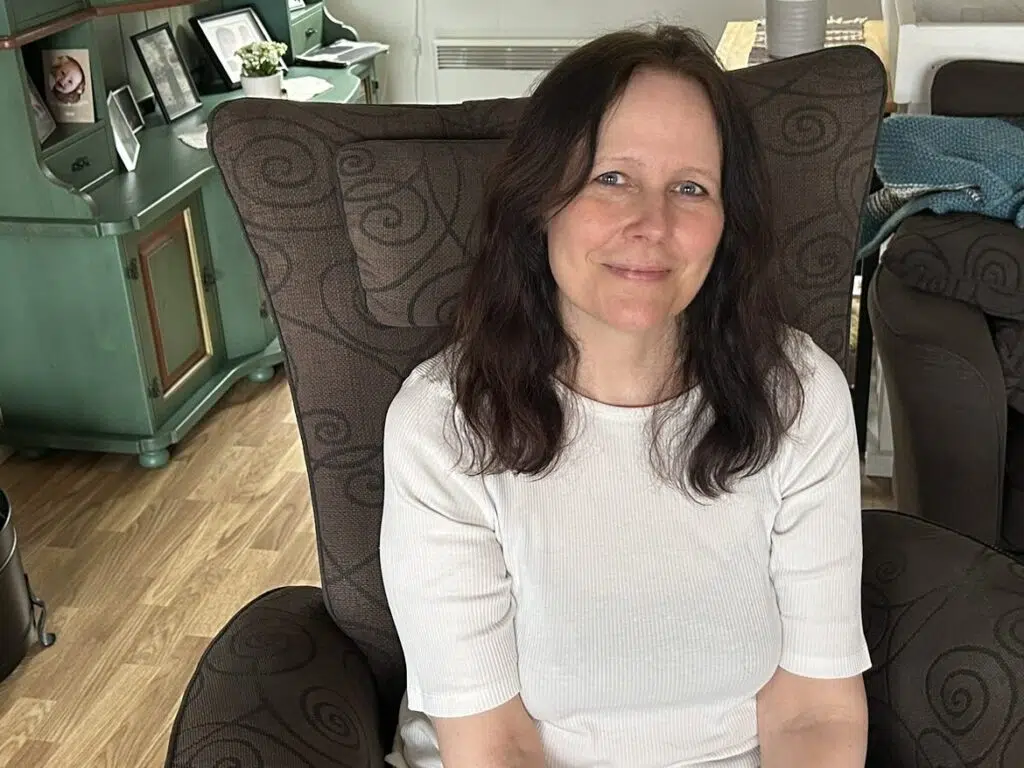 En kvinne med langt brunt hår sitter i en mønstret lenestol i en stue og smiler til kameraet. Det er et grønt skap og en brun sofa i bakgrunnen.