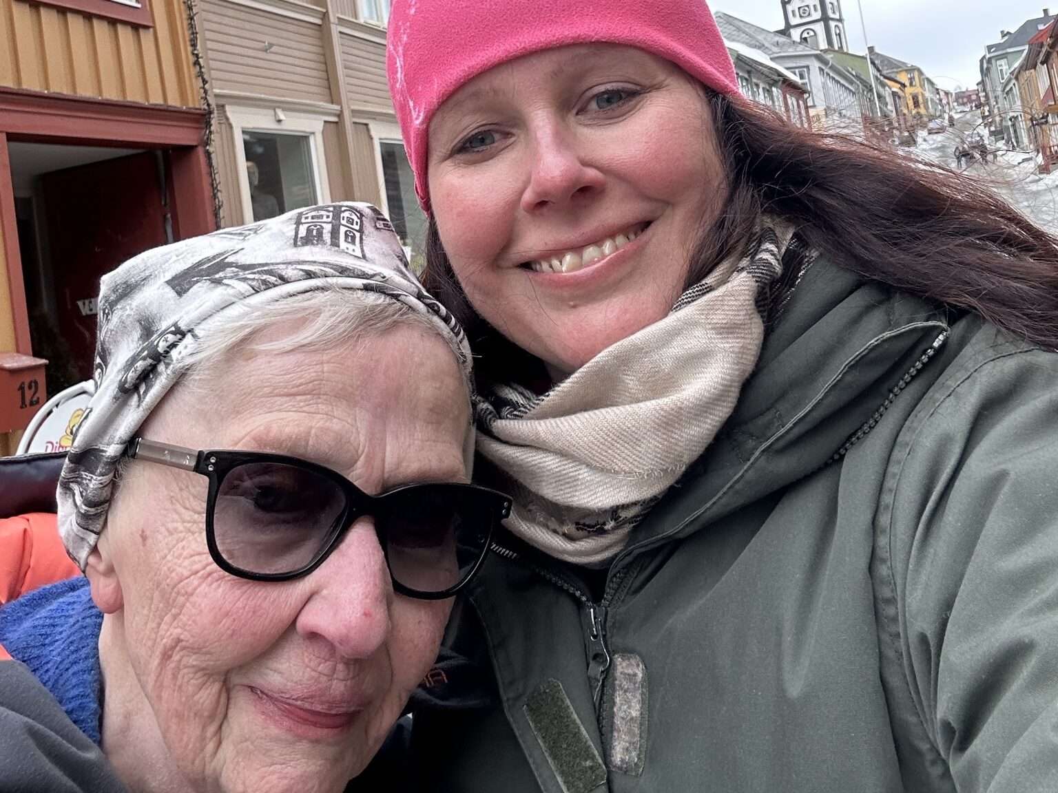 To kvinner i vinterklær og luer tar en selfie på en gate omgitt av trebygninger på en overskyet dag.