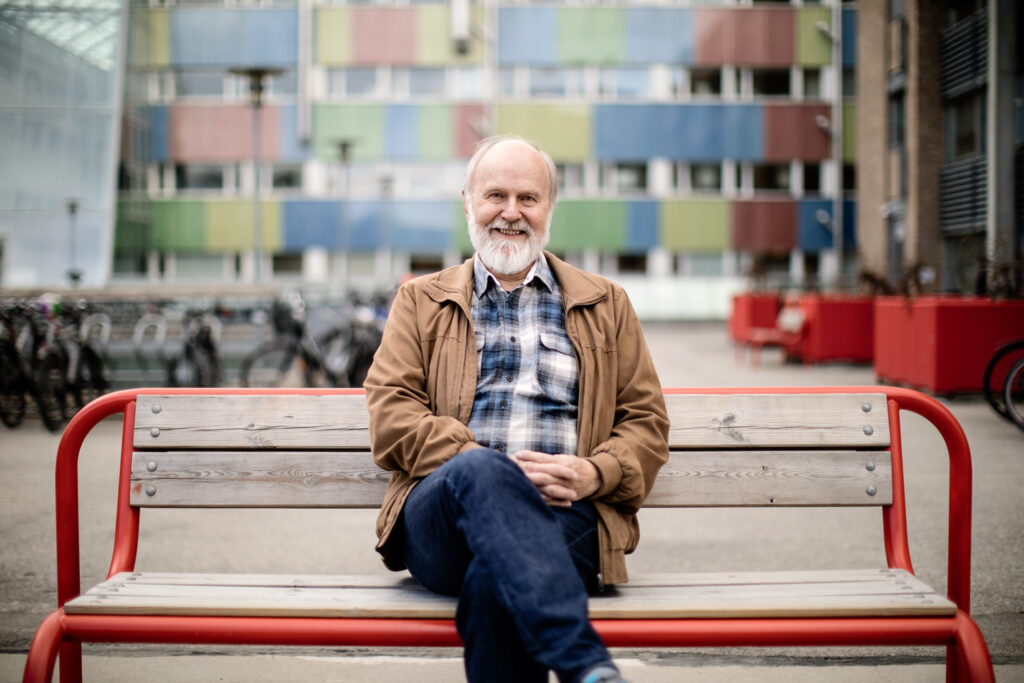 Mann som sitter på en parkbenk, iført en brun jakke og jeans, smiler. Bakgrunnen viser fargerik bygningsfasade og parkerte sykler.