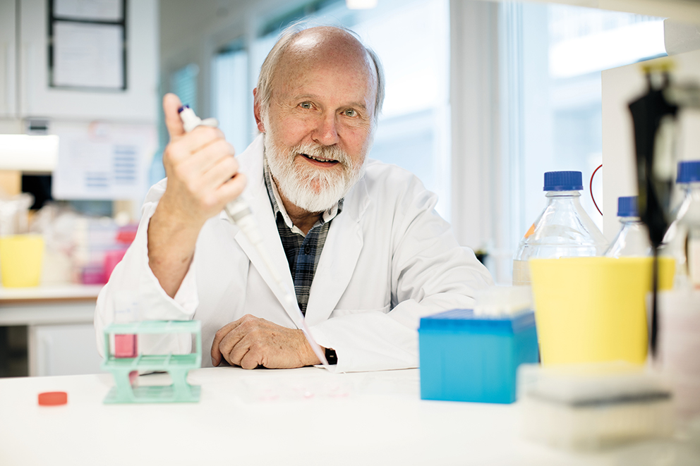 En eldre mann i laboratoriefrakk smiler mens han holder en pipette i laboratoriesetting med flasker og utstyr på bordet.