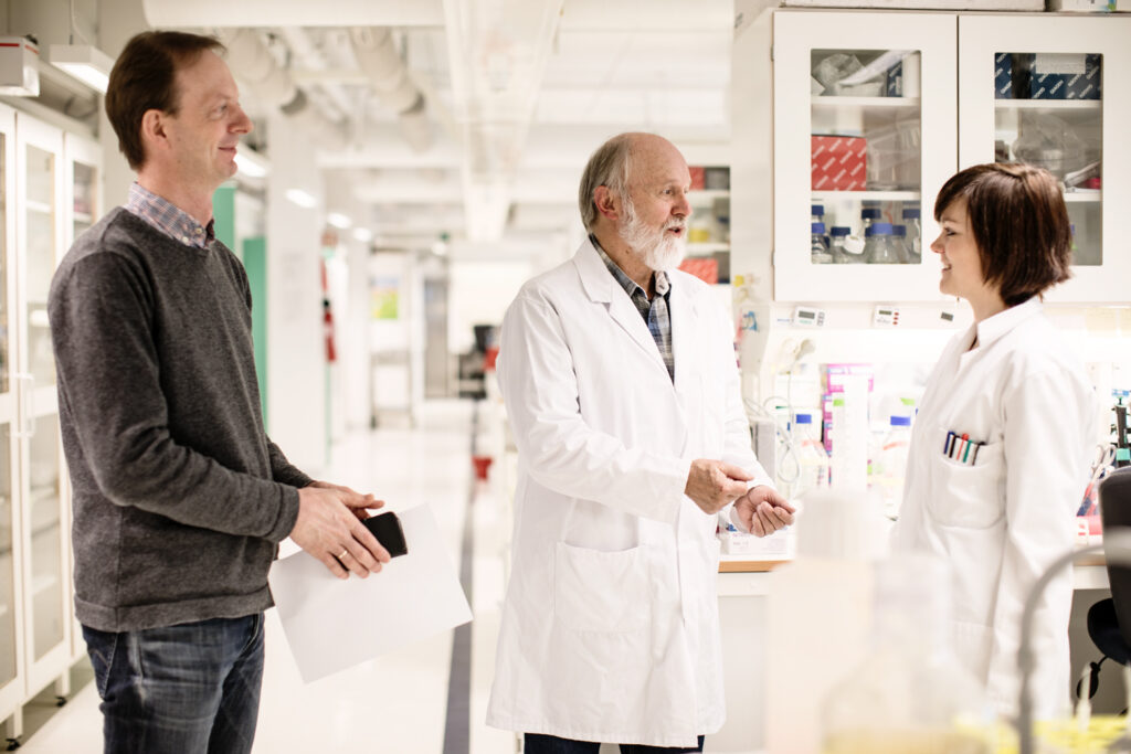 Tre personer i et laboratorium: en mann i en genser holder papirer, en annen i en labfrakk gester, og en kvinne i en labfrakk smiler. Hyller med laboratorieutstyr er i bakgrunnen.