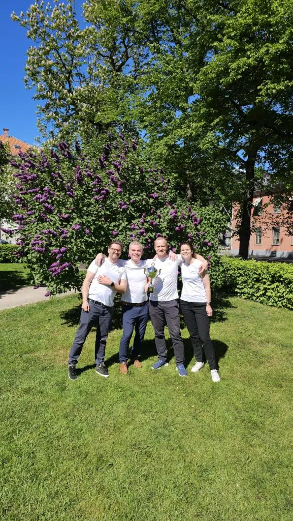 Fire personer i hvite skjorter står på gress foran en blomstrende busk og poserer sammen med et trofé etter Holmenkollstafetten på en solskinnsdag.