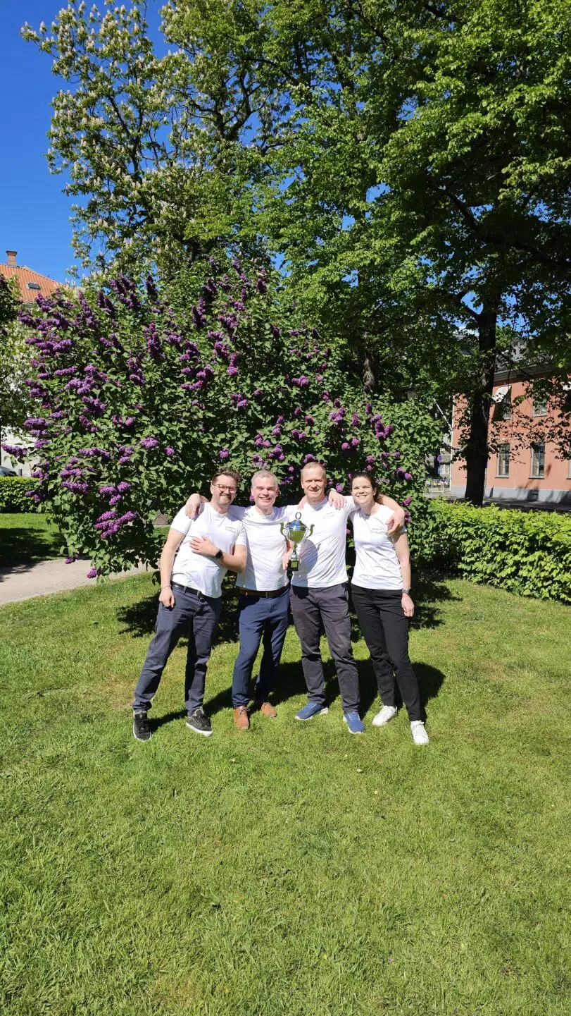 Fire personer i hvite skjorter står på gress foran en blomstrende busk og poserer sammen med et trofé etter Holmenkollstafetten på en solskinnsdag.