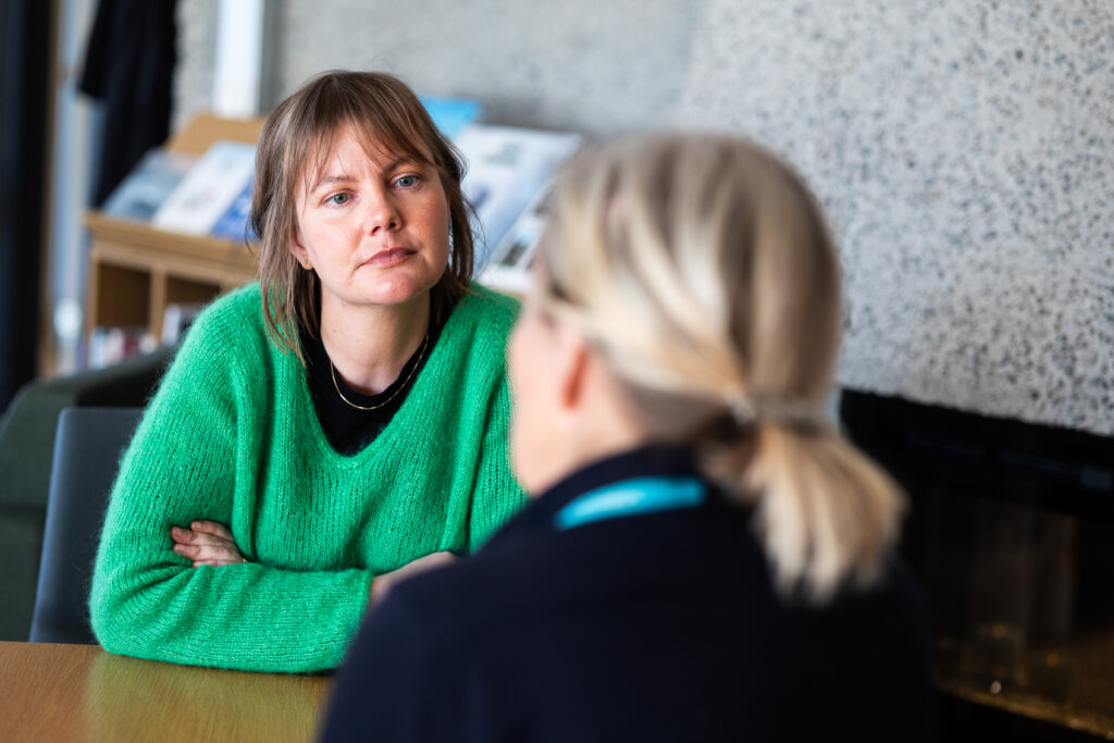 To personer sitter ved et bord innendørs og har en alvorlig samtale. Personen som står overfor kameraet har på seg en grønn genser og lytter oppmerksomt.