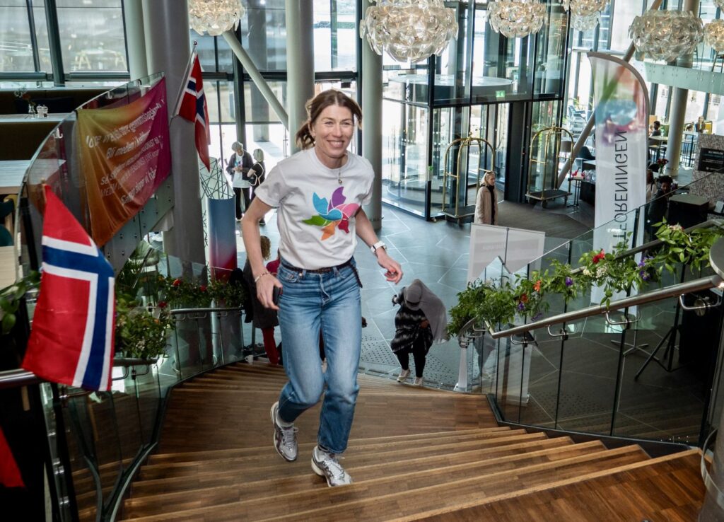 En kvinne i hvit t-skjorte og jeans går opp en tretrapp inne i en moderne bygning dekorert med norske flagg og bannere.