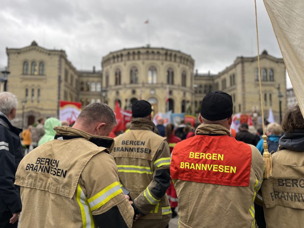 Flere brannmenn med «Bergen Brannvesen» på jakkene sine står foran en folkemengde i nærheten av en utsmykket bygning på en overskyet dag.