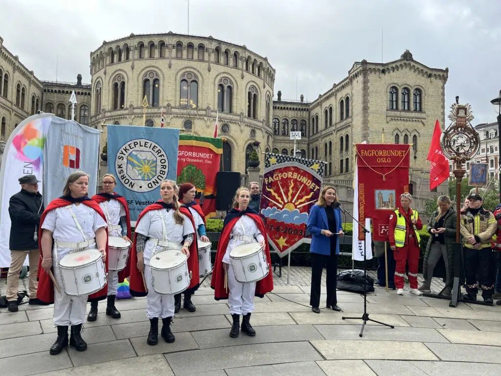 En gruppe uniformerte trommeslagere står foran bannere og en historisk bygning, med en kvinne som snakker ved en mikrofon og flere personer som observerer.