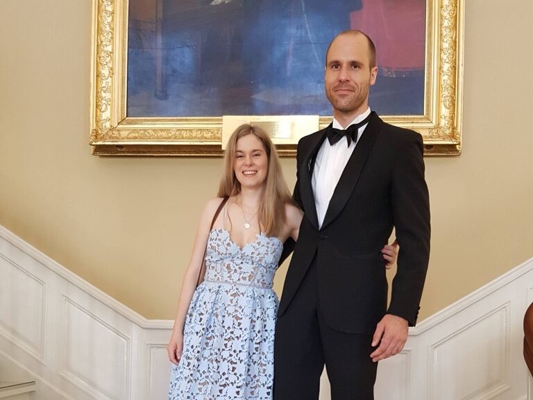 En kvinne i en lyseblå blondekjole og en mann i en svart smoking står sammen på en innendørs trapp og poserer for et formelt bilde.