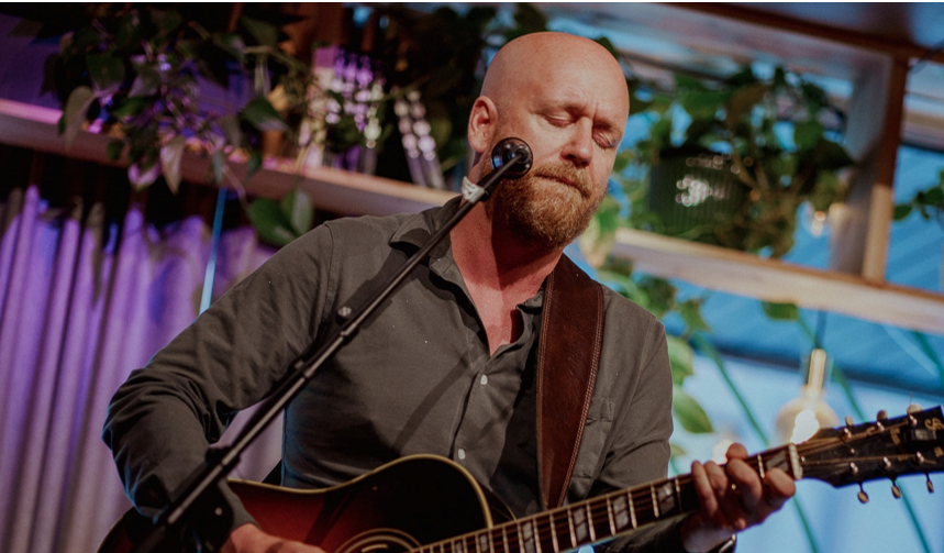 En mann med skjegg spiller akustisk gitar og synger inn i en mikrofon på scenen, med planter og lys i bakgrunnen.