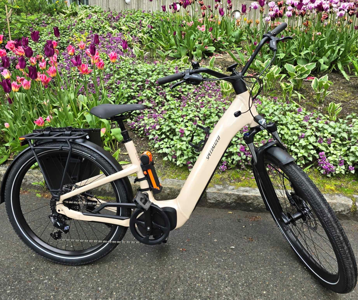 En beige Specialized elsykkel står parkert på en asfaltert sti ved siden av et hagebed med blomstrende tulipaner og grøntområder.