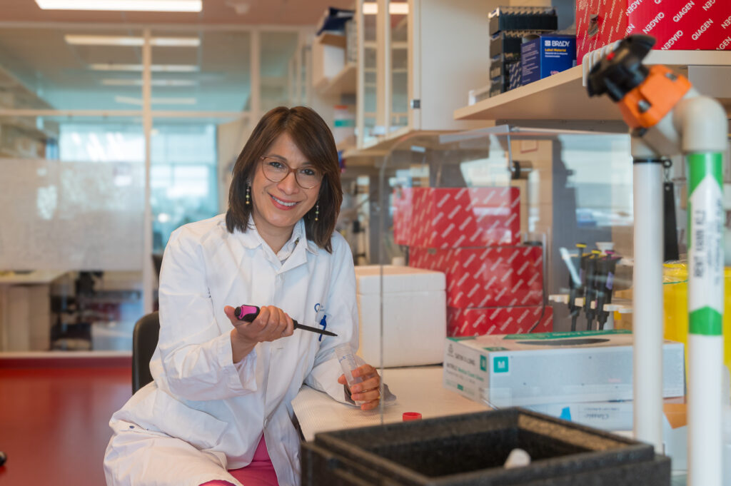 En forsker i laboratoriefrakk sitter ved en laboratoriebenk, holder en pipette og smiler til kameraet. Laboratorieutstyr og -forsyninger er synlige i bakgrunnen.