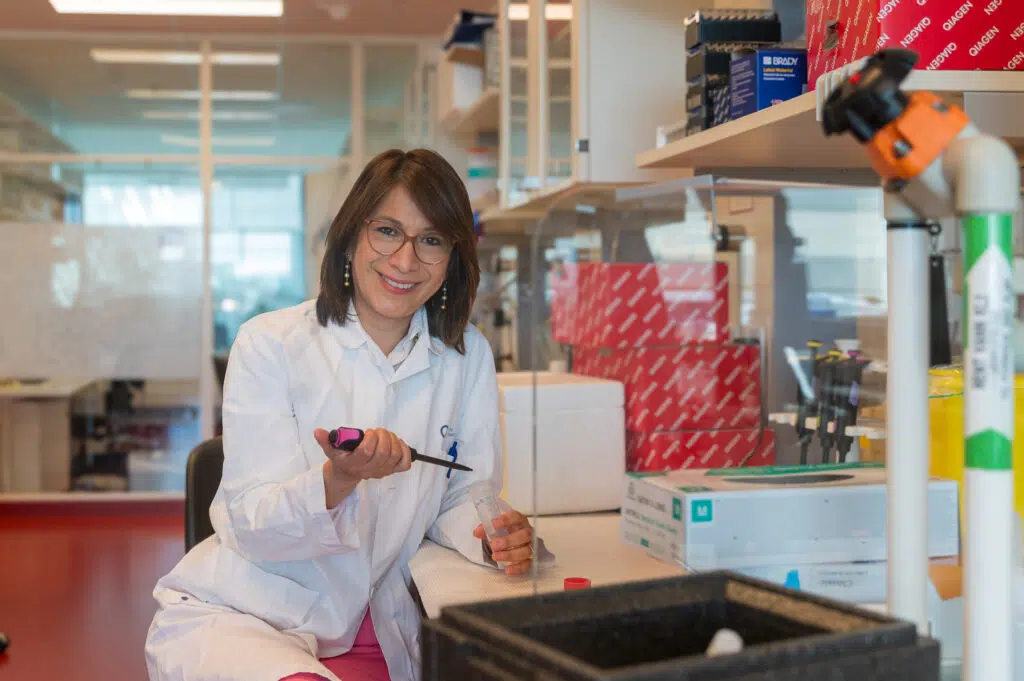 En forsker i laboratoriefrakk sitter ved en laboratoriebenk, holder en pipette og smiler til kameraet. Laboratorieutstyr og -forsyninger er synlige i bakgrunnen.