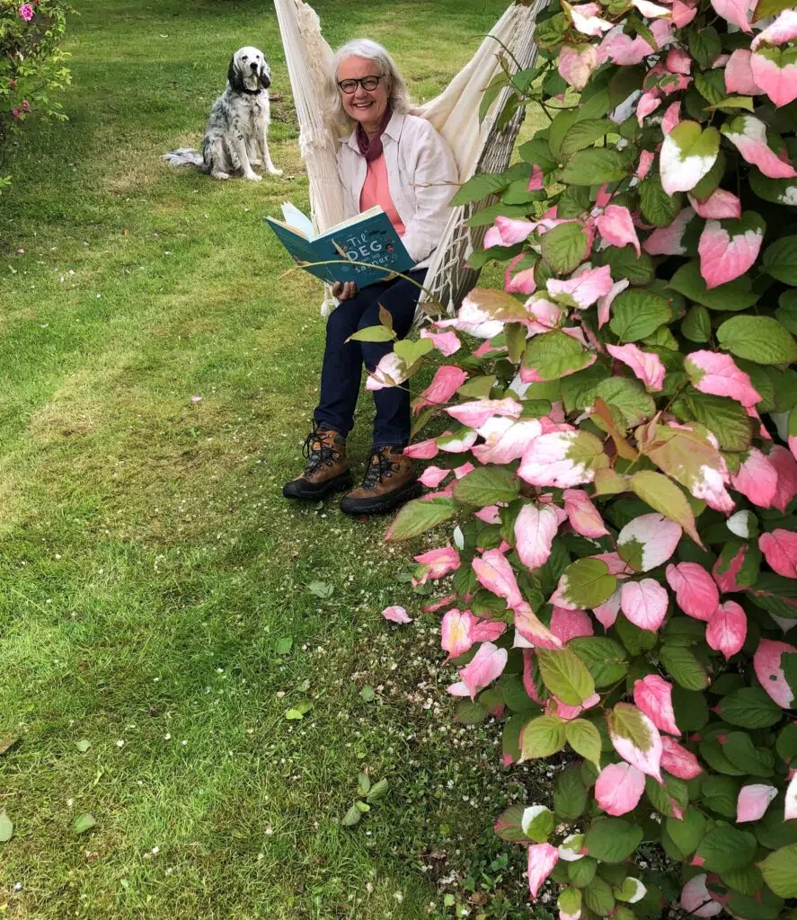 En kvinne sitter i en hengekøye og leser en bok i en hage, med en hund som sitter i gresset like ved og rosa blomster i forgrunnen.