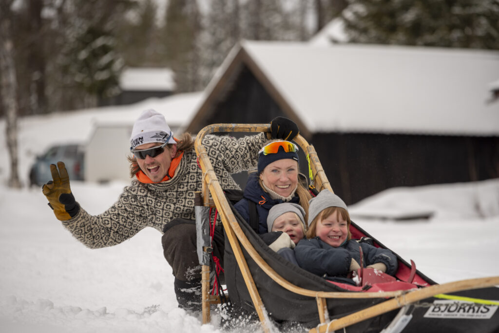 En mann og kvinne med to barn kjører hundeslede på snø og demonstrerer samarbeid mens de nyter eventyret sammen. Mannen smiler og lager et fredstegn, med snødekte hytter og trær i bakgrunnen.