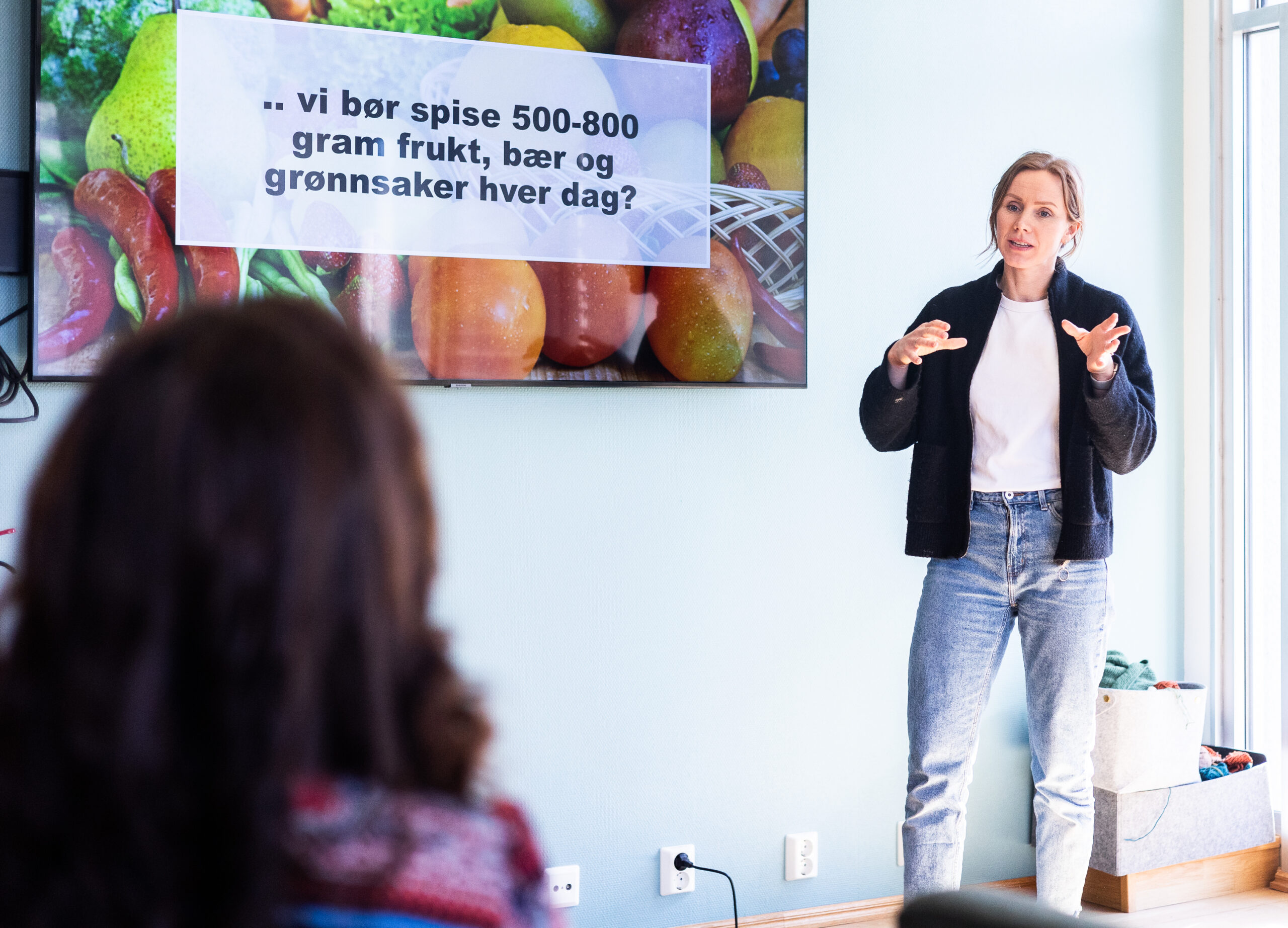 En kvinne holder et foredrag om ernæring, stående ved en skjerm som viser et budskap på norsk om å spise 500-800 gram frukt, bær og grønnsaker daglig.