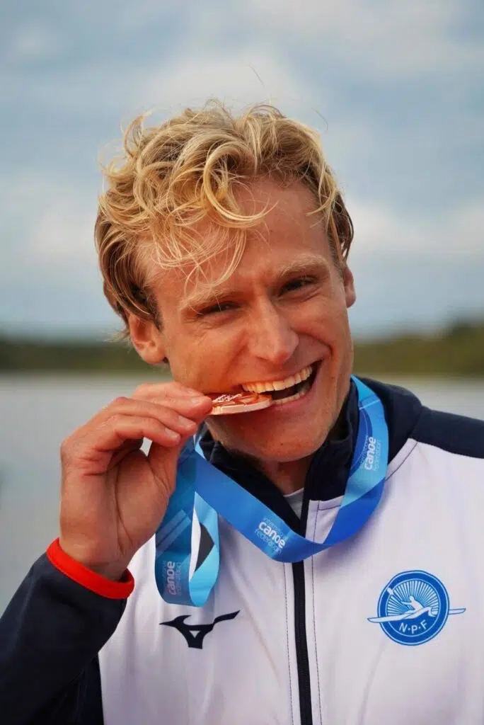 En mann iført sportsjakke og blått bånd med medalje biter på medaljen mens han smiler utendørs.