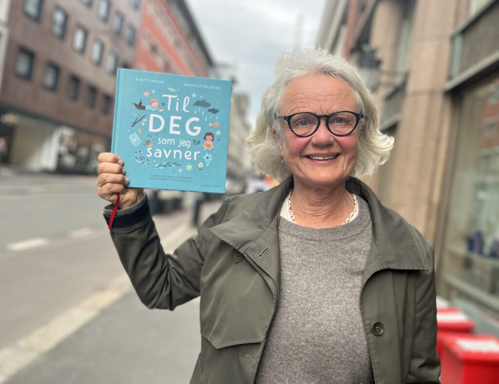 En eldre kvinne med briller smiler mens hun holder opp en bok med tittelen "Til deg som jeg savner" i en bygate.