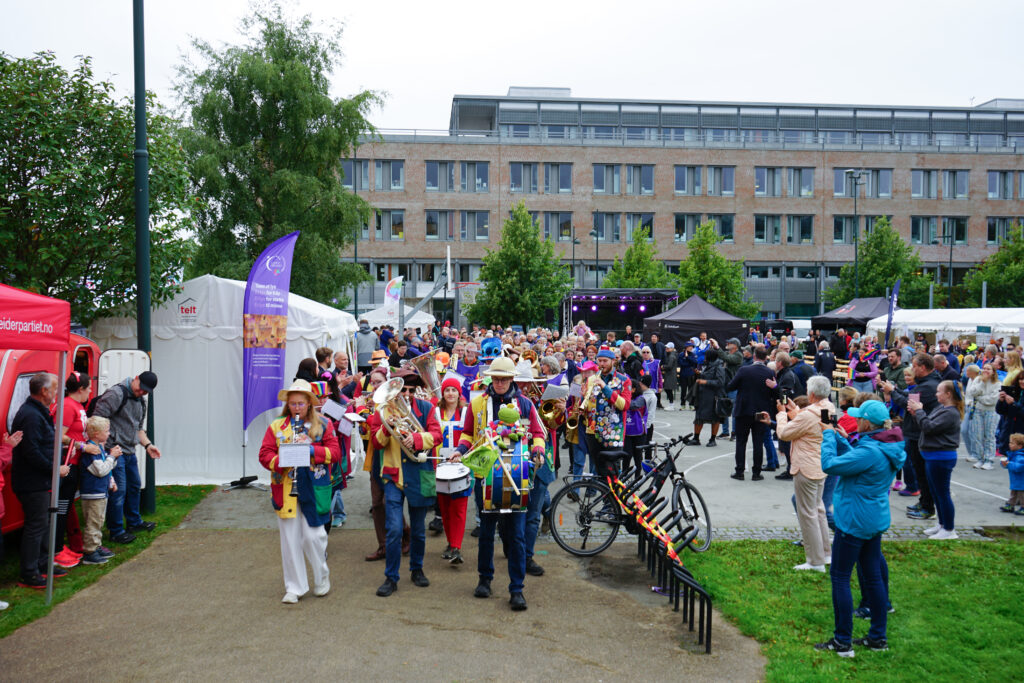 Et kostymekledd marsjorkester spiller instrumenter på et utendørsarrangement; folk ser på, og telt og en moderne bygning er synlige i bakgrunnen.