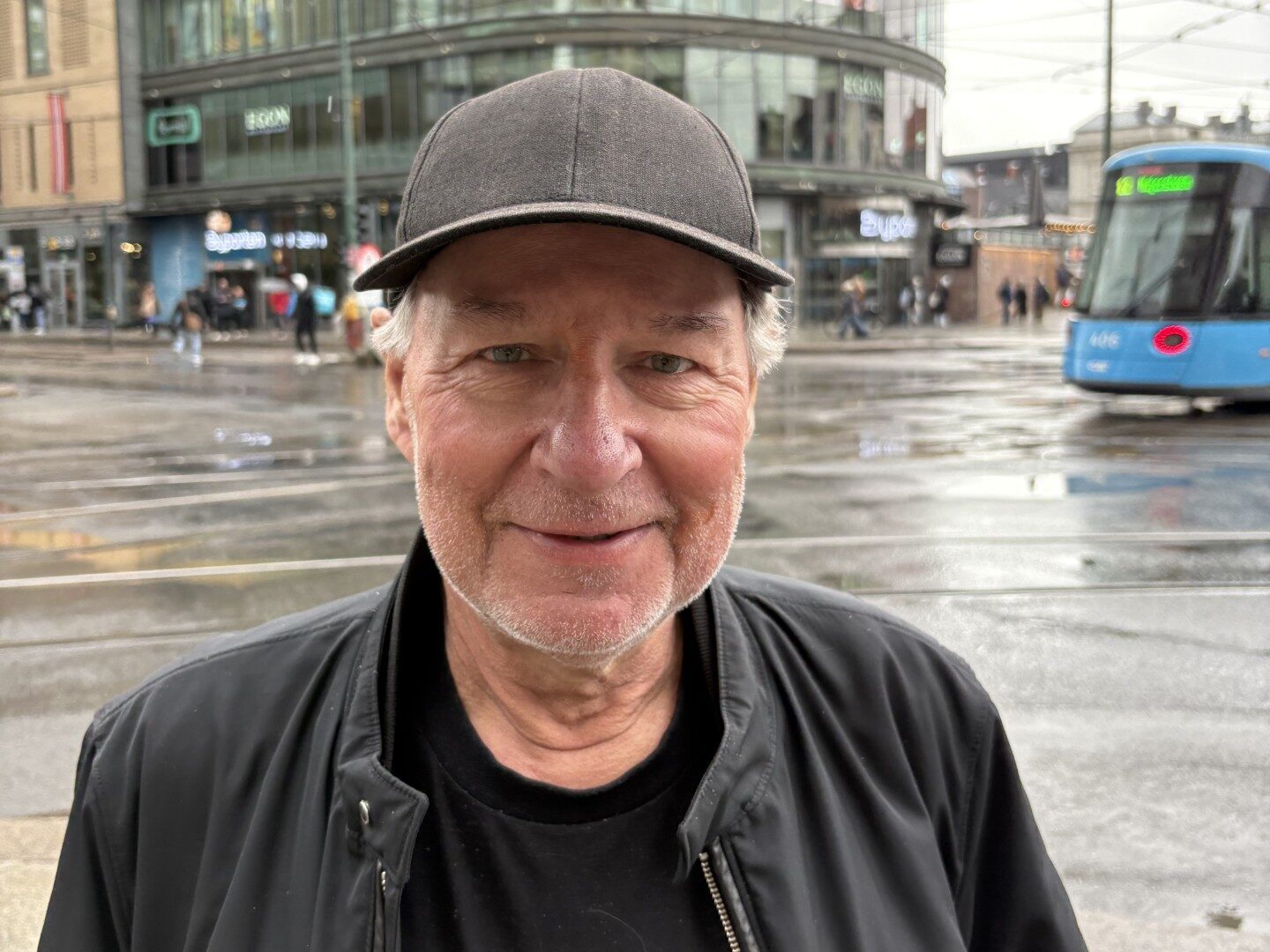 En mann i svart lue og jakke står utenfor på en våt gate med Kjøpesenteret i Oslo og en blå trikk synlig i bakgrunnen.