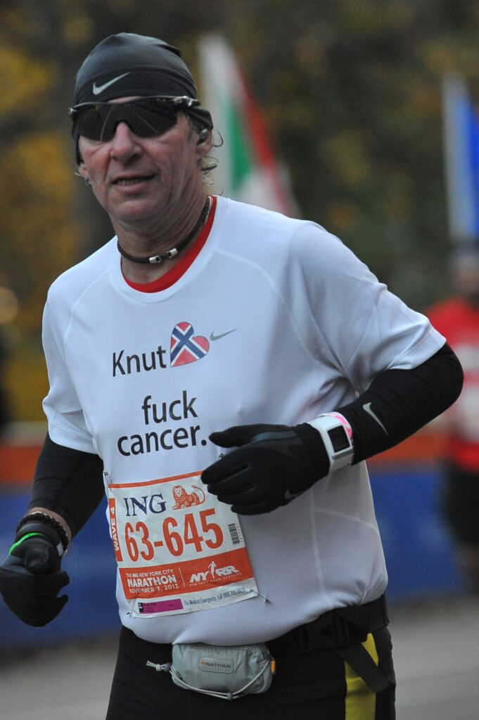 En løper med solbriller og en «Knut fuck cancer»-skjorte deltar i et maraton, med en startnummer 63–645 synlig på livet.