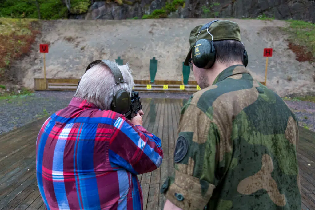 En eldre person i rutete skjorte sikter en pistol mot mål på en utendørs skytebane, ledsaget av en person i kamuflasjeuniform og hørselsvern.
