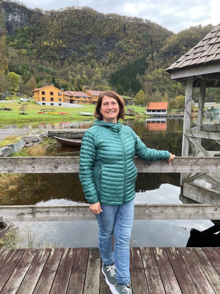 En kvinne i en grønn dunjakke står og smiler på en trebrygge, med en innsjø, et gresskledd område og fjell i bakgrunnen.