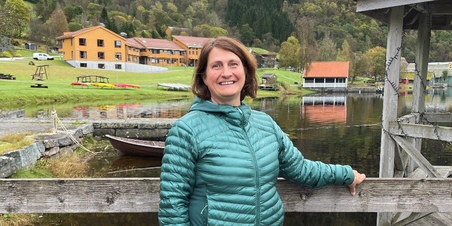 En kvinne i en grønn dunjakke står og smiler på en trebrygge, med en innsjø, et gresskledd område og fjell i bakgrunnen.