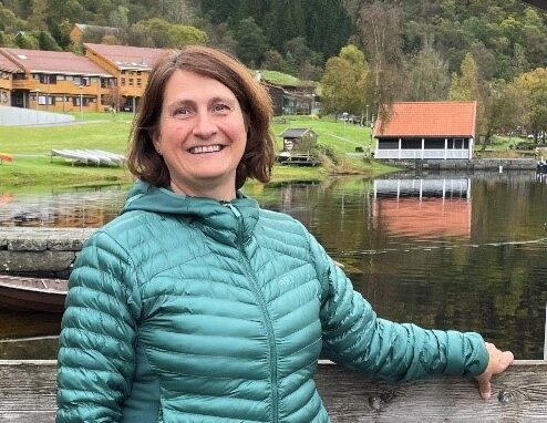 En kvinne i en blågrønn jakke står på en trebrygge ved en innsjø, med hytter, grønne åser og trær i bakgrunnen.