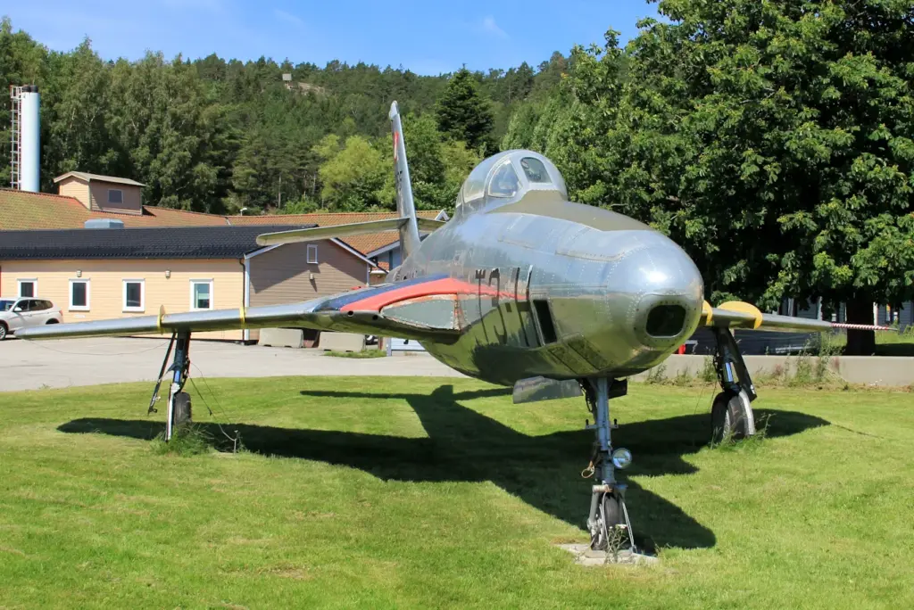 Et sølvfarget vintagejagerfly er utstilt på et gresskledd område i nærheten av bygninger og trær under en klar himmel.