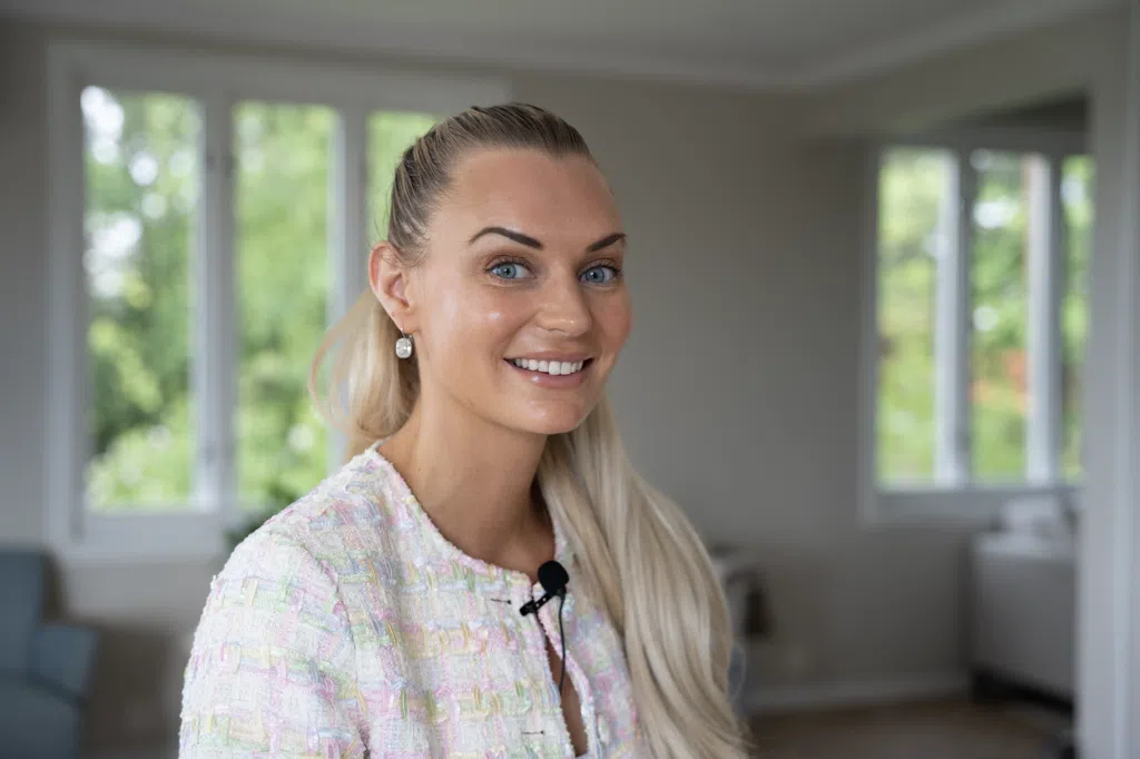 En kvinne med langt blondt hår, iført en lys, teksturert blazer og en mikrofon med krage, smiler i et lyst rom med store vinduer.