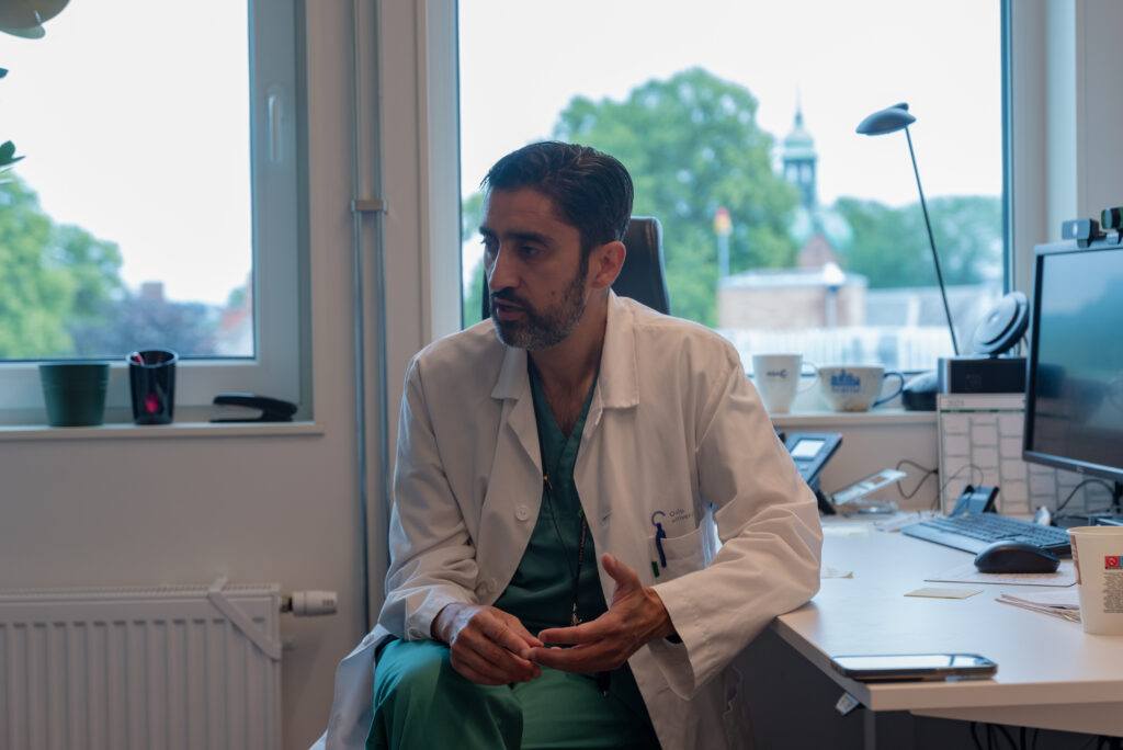 En mann i hvit laboratoriefrakk sitter og snakker på et kontor, omgitt av medisinsk utstyr og papirarbeid, med et vindu i bakgrunnen.