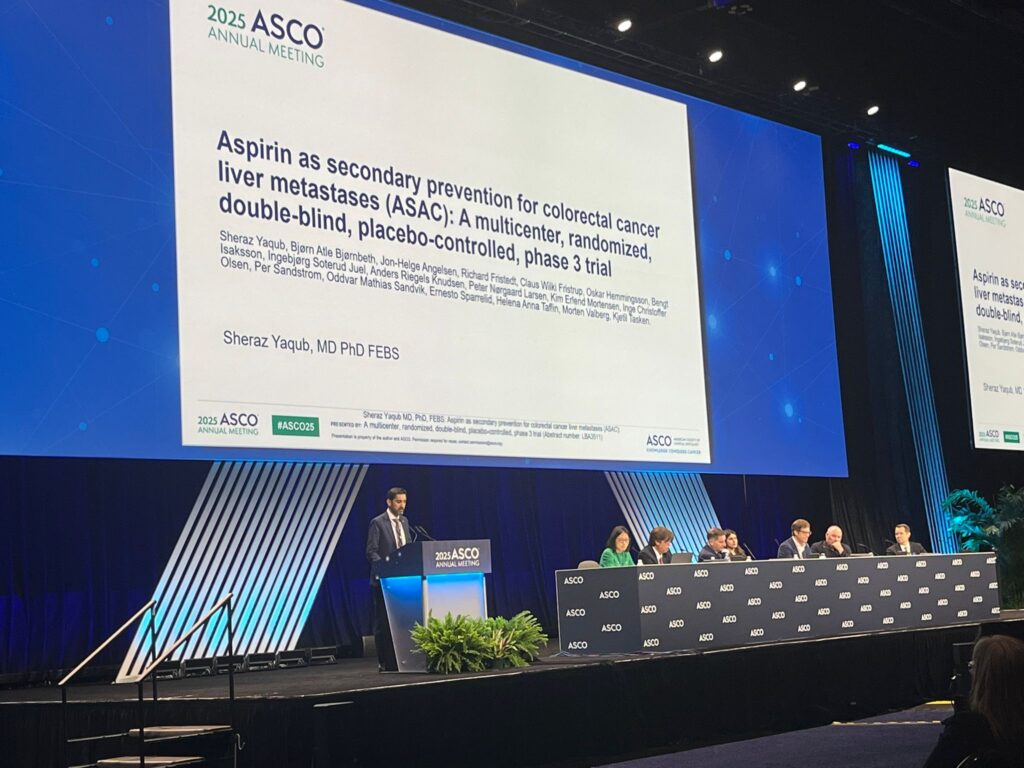En foredragsholder presenterer en klinisk studie med aspirin for behandling av levermetastaser ved kolorektal kreft på ASCOs årsmøte i 2025, mens et panel sitter ved et langbord på scenen.
