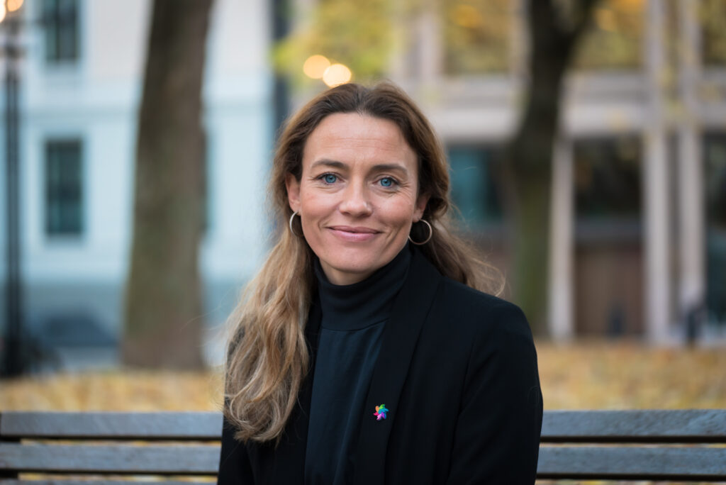 En kvinne med langt, lysebrunt hår, iført svart jakke og polotrøye, sitter på en benk utendørs med trær og bygninger i bakgrunnen.