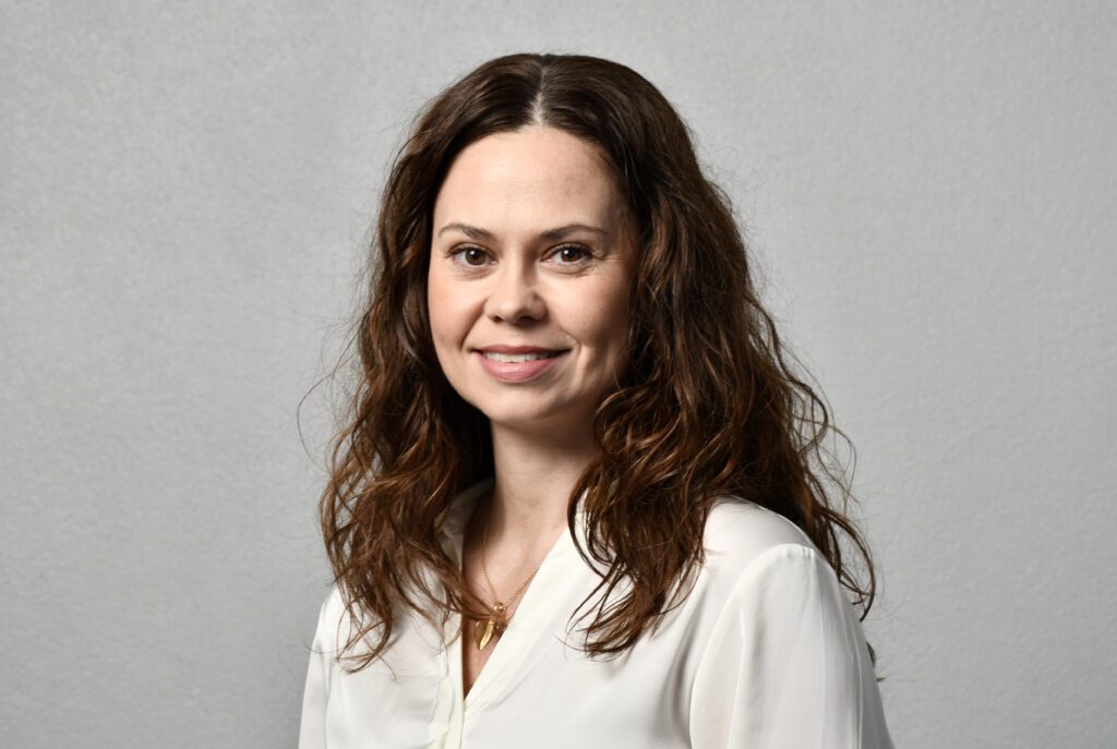 Kvinne med bølget, brunt hår og hvit bluse, smilende, avbildet mot en vanlig, lysegrå bakgrunn.