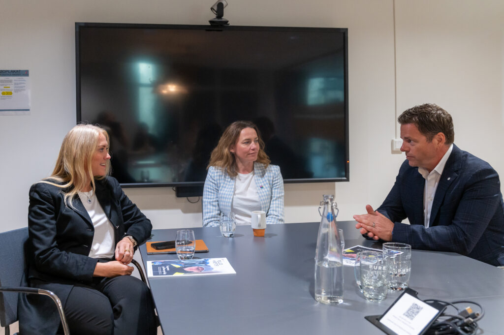 Tre personer i forretningsantrekk sitter rundt et konferansebord og diskuterer, med papirer, briller og en stor skjerm synlig i bakgrunnen.