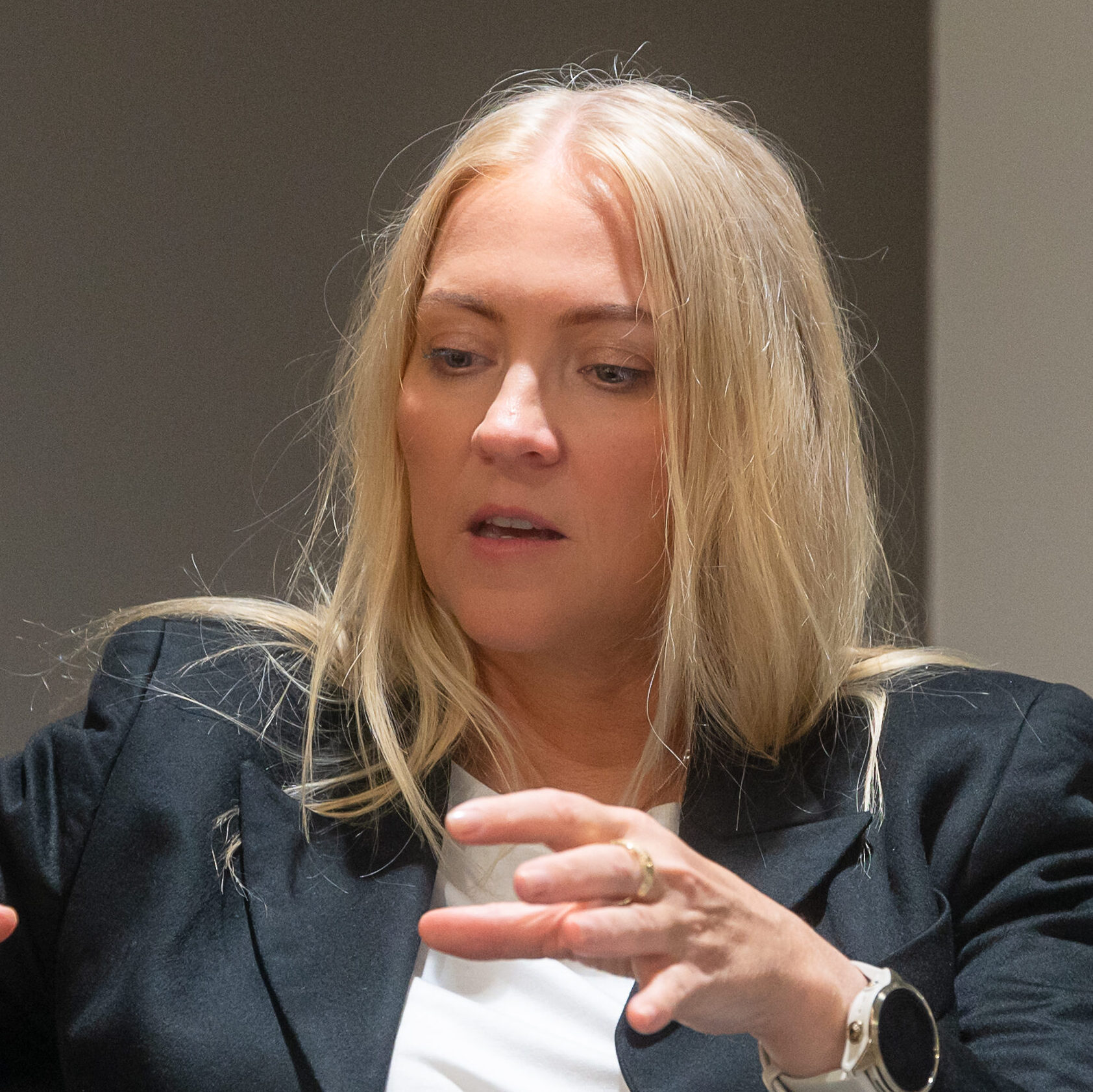 En kvinne med langt, blondt hår og svart blazer gestikulerer med hendene mens hun snakker ved et bord med flere tomme glass foran seg.