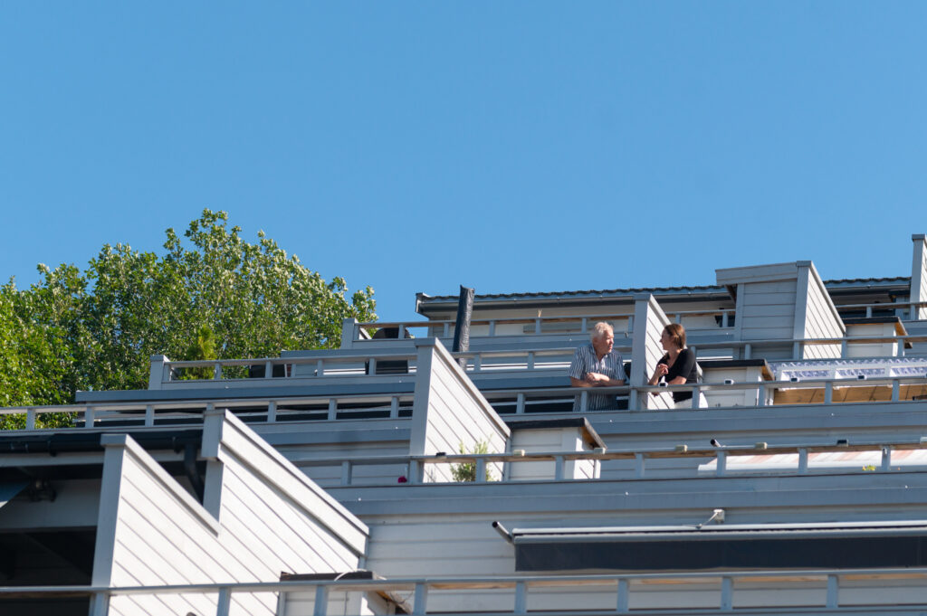 To personer står og snakker sammen på en balkong i en moderne bygning med flere etasjer og klar blå himmel i bakgrunnen.