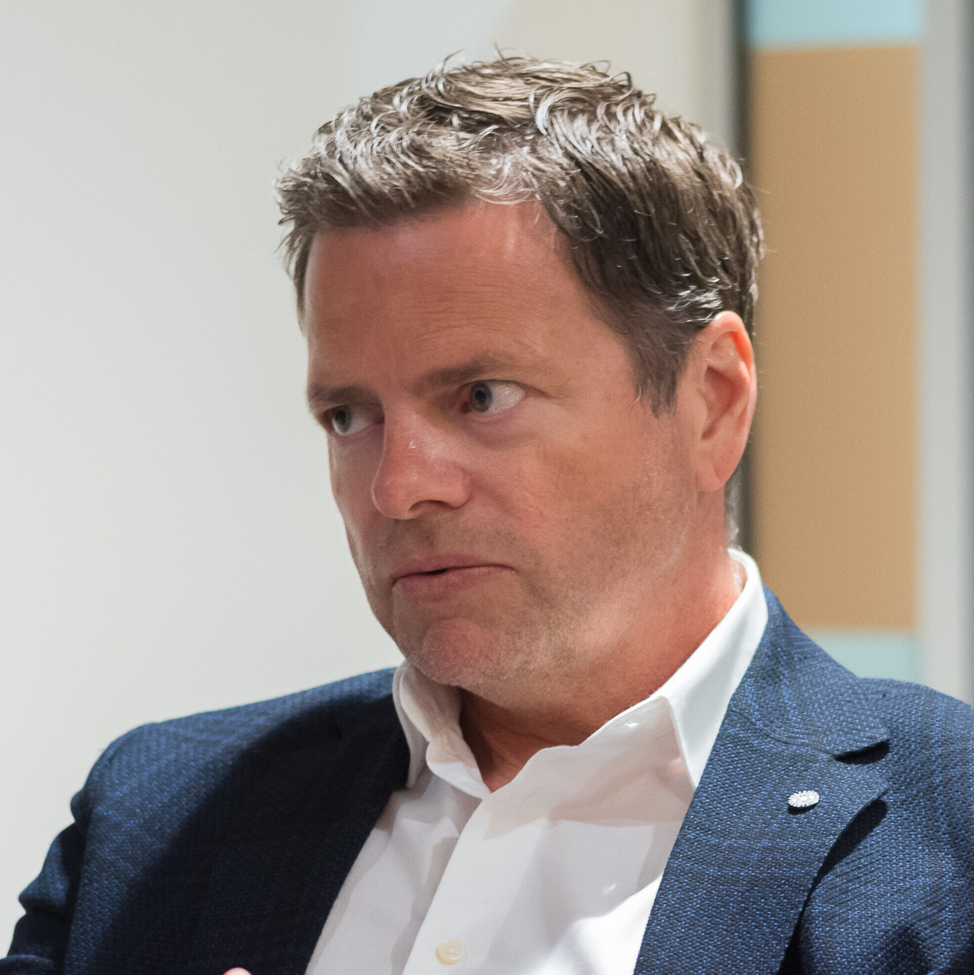 En mann i blå blazer og hvit skjorte sitter ved et bord og gestikulerer med hendene under en samtale i et kontormiljø.