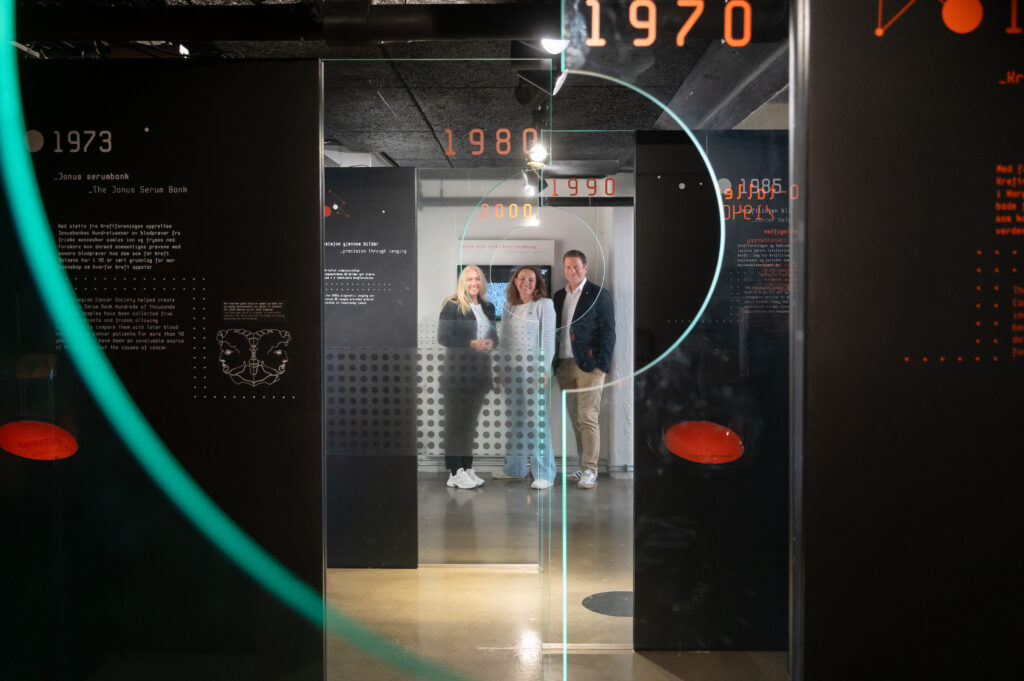 Tre personer står sammen i en moderne museumsutstilling, omgitt av mørke paneler med tekst, tall og grafikk som forteller om 1970- og 1980-tallet.