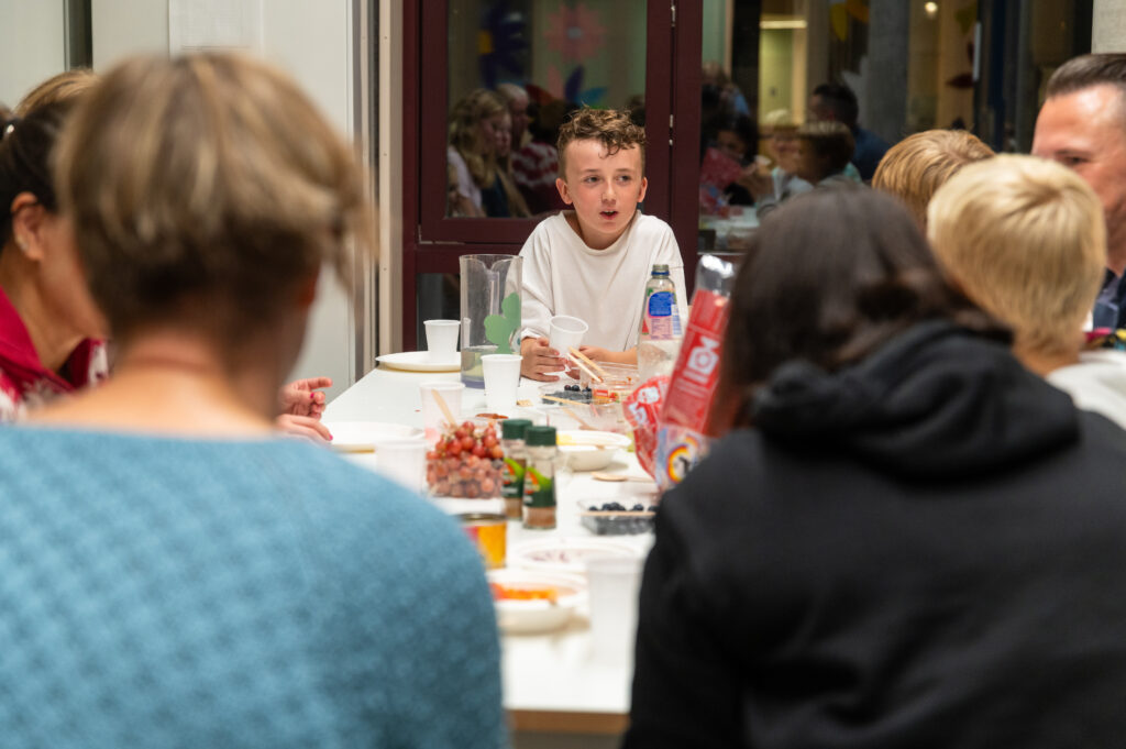 En gruppe mennesker sitter rundt et bord med mat, mens en gutt i hvit skjorte snakker og de andre lytter.